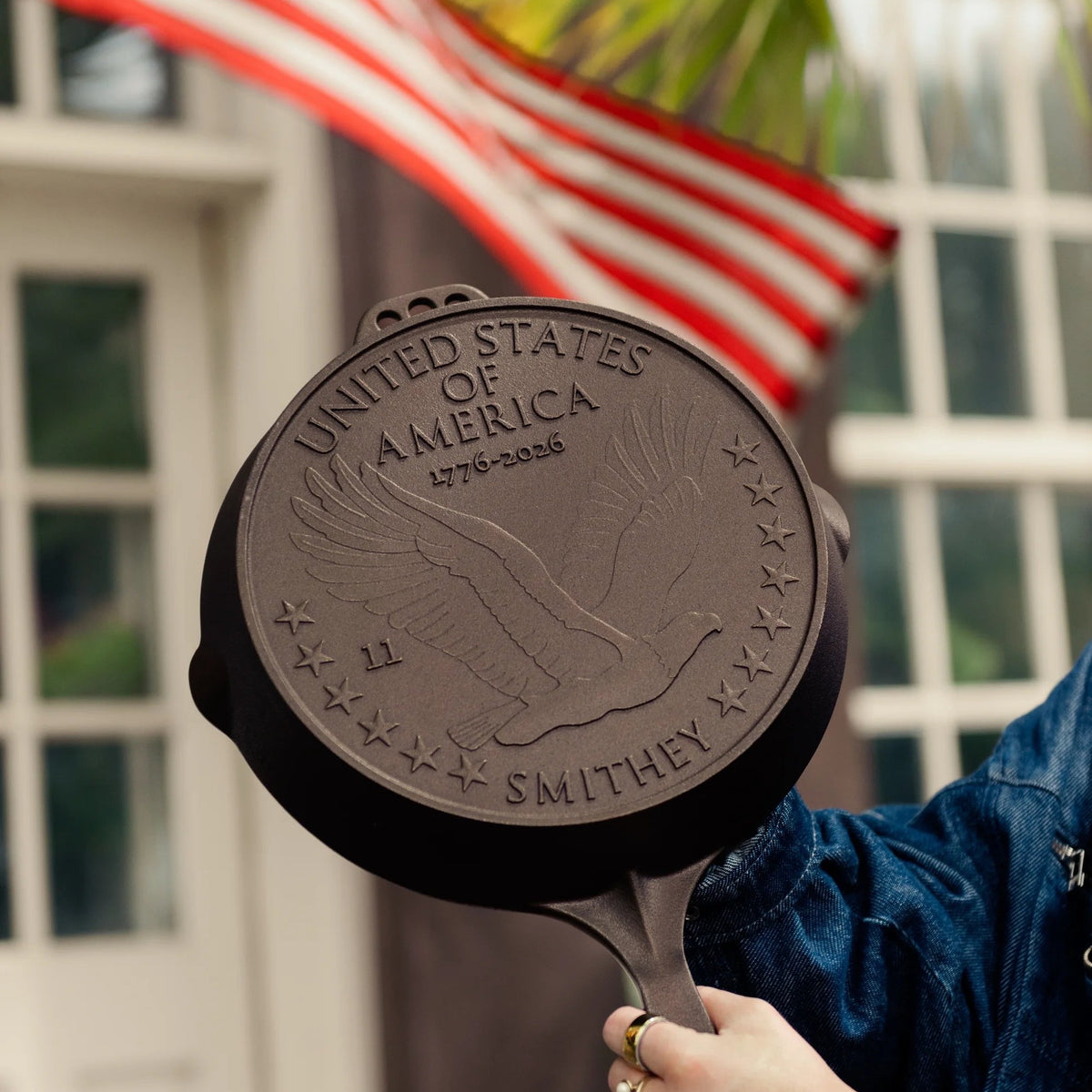 Hand holding the No. 11 American Anniversary Skillet up to show its embossed eagle and 1776–2026, with an American flag behind it.
